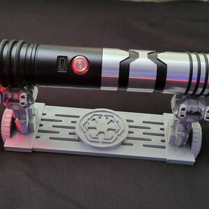 May include: A black and silver lightsaber with a red blade is displayed on a gray stand with a black and white symbol. The lightsaber has a black handle with a silver accent and a black and silver blade. The stand has a black and white symbol on the front.