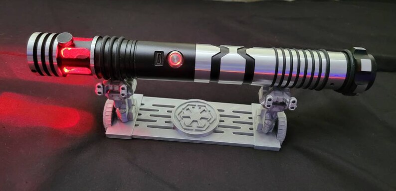 May include: A black and silver lightsaber with a red blade is displayed on a gray stand with a black and white symbol. The lightsaber has a black handle with a silver accent and a black and silver blade. The stand has a black and white symbol on the front.
