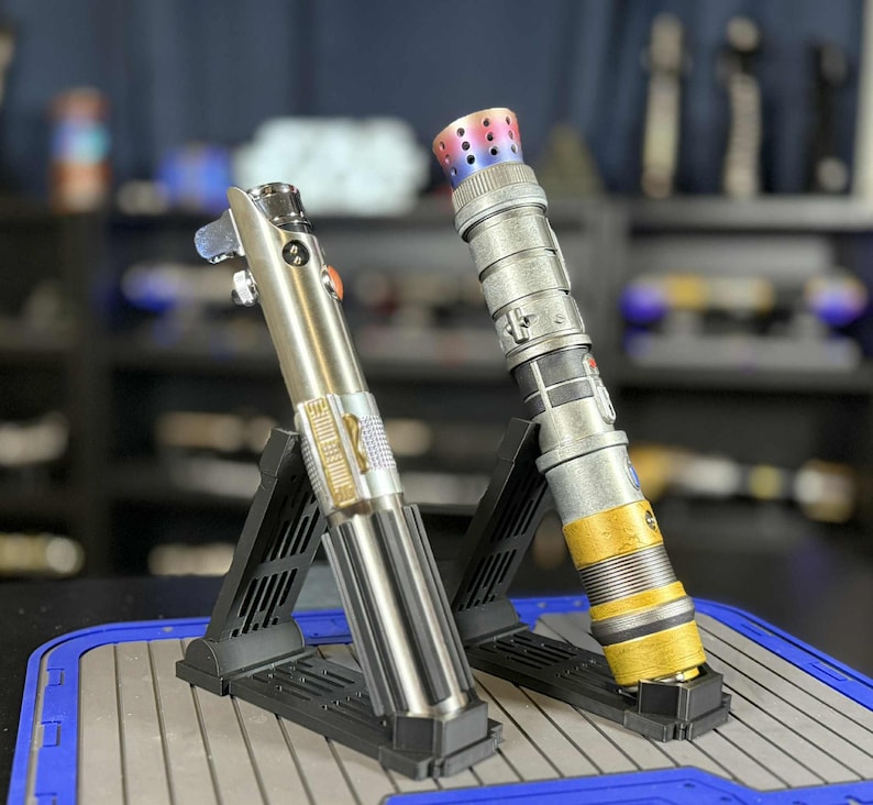K&ouml;nnte beinhalten: Zwei metallische Lichtschwerter, eines silberfarben und eines mit silber-goldener Oberfl&auml;che, werden auf schwarzen St&auml;ndern pr&auml;sentiert. Die Lichtschwerter weisen komplizierte Details auf und heben sich vor einem verschwommenen Hintergrund anderer Sammlerst&uuml;cke ab.
