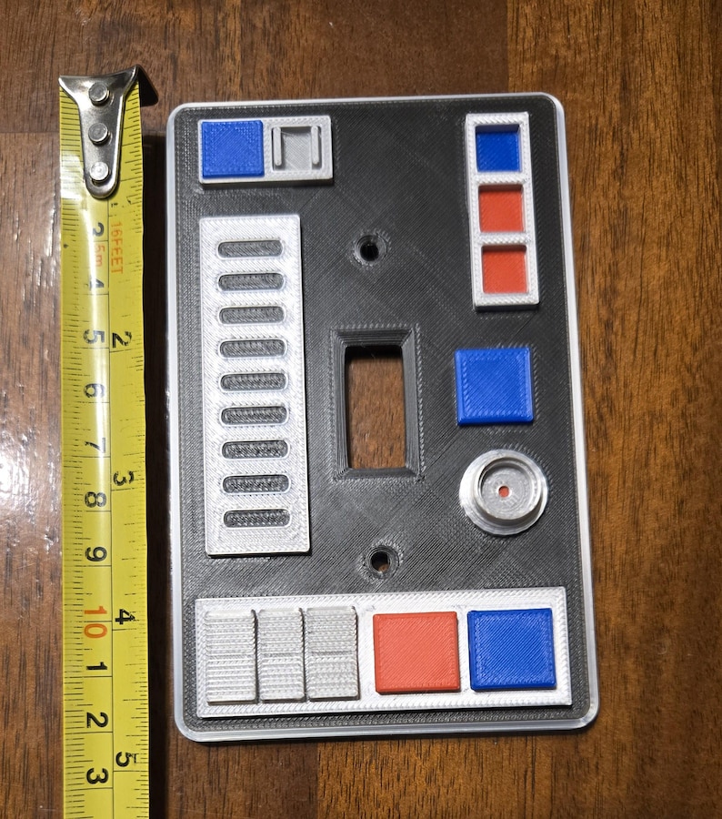 Op de afbeelding: Een zwarte lichtschakelaar-afdekplaat met een zilveren en grijs ontwerp. De afdekplaat heeft verschillende knoppen, waaronder rode, blauwe en zilveren. De afdekplaat is 10 cm hoog.