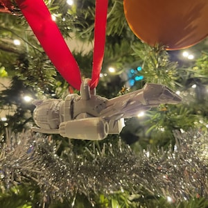 May include: A white 3D printed spaceship ornament hanging from a red ribbon on a Christmas tree. The ornament is a detailed model of a spaceship with a pointed nose and two engines.