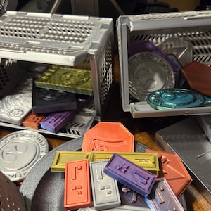 May include: Two silver metal storage boxes with open lids, revealing various colorful plastic tokens and chips. The boxes are on a table with a silver metal surface.