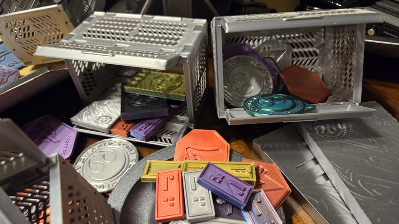 May include: Two silver metal storage boxes with open lids, revealing various colorful plastic tokens and chips. The boxes are on a table with a silver metal surface.