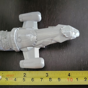 May include: A gray 3D printed model of a spaceship, approximately 10 inches long, with a detailed design and a textured surface. The model is sitting on a table next to a measuring tape.