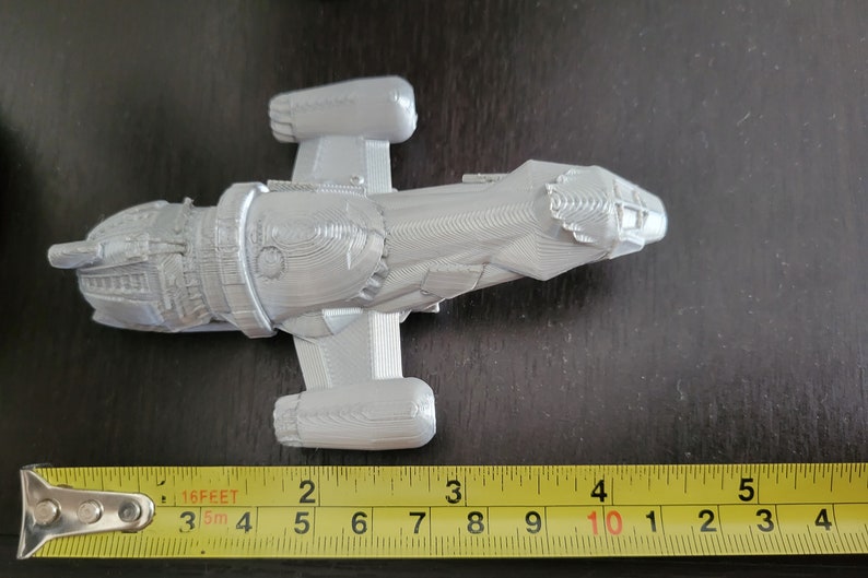 May include: A gray 3D printed model of a spaceship, approximately 10 inches long, with a detailed design and a textured surface. The model is sitting on a table next to a measuring tape.