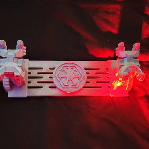 May include: A white 3D printed Star Wars themed phone stand with two TIE fighter models on either side of a circular Imperial symbol. The stand is lit with red LED lights.