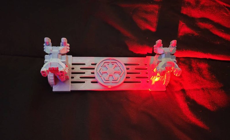 May include: A white 3D printed Star Wars themed phone stand with two TIE fighter models on either side of a circular Imperial symbol. The stand is lit with red LED lights.