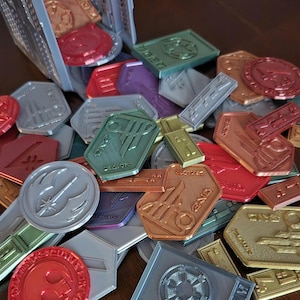 May include: A collection of colorful, metallic, and silver tokens with various symbols and designs. The tokens are shaped like circles, squares, and hexagons. Some of the tokens have text on them, including "MEDIC", "REBEL ALLIANCE", and "GALACTIC EMPIRE".