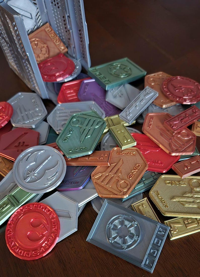 May include: A collection of colorful, metallic, and silver tokens with various symbols and designs. The tokens are shaped like circles, squares, and hexagons. Some of the tokens have text on them, including "MEDIC", "REBEL ALLIANCE", and "GALACTIC EMPIRE".