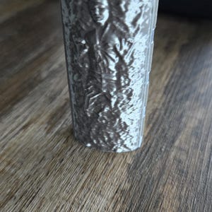 May include: A silver-colored lighter with a red ignition button. The lighter's body features a detailed relief of a person. The lighter is on a wooden surface.