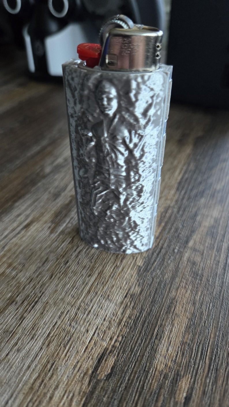 May include: A silver-colored lighter with a red ignition button. The lighter's body features a detailed relief of a person. The lighter is on a wooden surface.