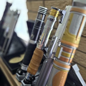 K&ouml;nnte beinhalten: Eine Sammlung von Lichtschwertern in verschiedenen Designs, die auf einem Holzregal ausgestellt sind. Die Lichtschwerter weisen metallische und farbige Akzente auf, darunter Silber, Gold und Schwarz. Jedes Schwert ruht in einem individuellen St&auml;nder vor einer Backsteinmauer.