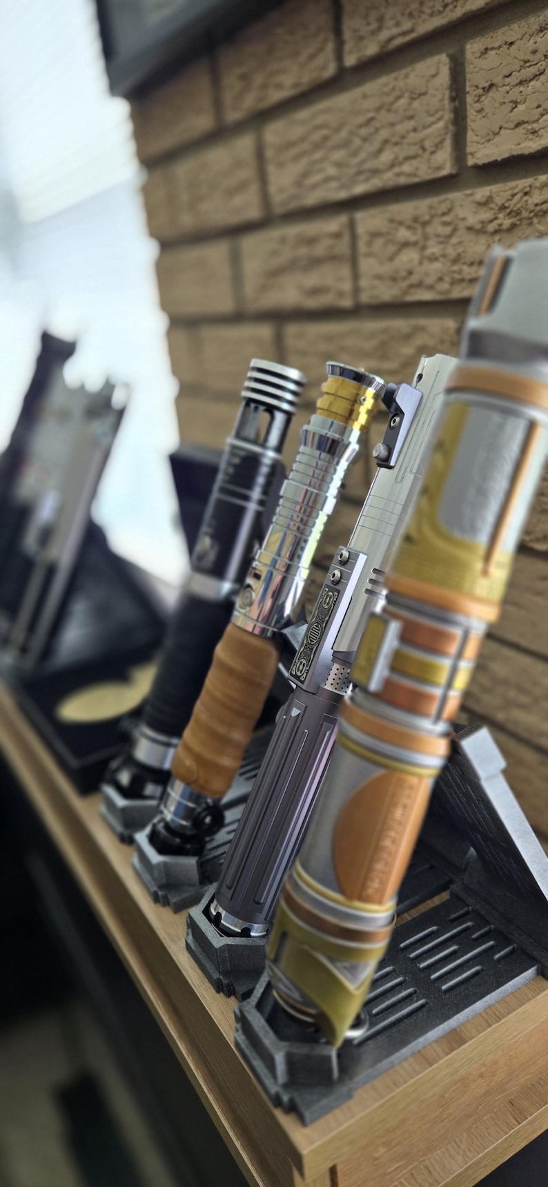 K&ouml;nnte beinhalten: Eine Sammlung von Lichtschwertern in verschiedenen Designs, die auf einem Holzregal ausgestellt sind. Die Lichtschwerter weisen metallische und farbige Akzente auf, darunter Silber, Gold und Schwarz. Jedes Schwert ruht in einem individuellen St&auml;nder vor einer Backsteinmauer.