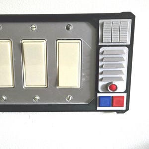 May include: A custom light switch cover with a silver faceplate and black trim. It features three light switches, a series of small rectangular and square details, and a red and blue button. The design has a retro-futuristic aesthetic.