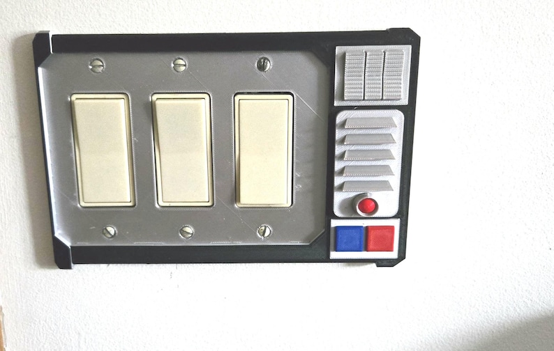May include: A custom light switch cover with a silver faceplate and black trim. It features three light switches, a series of small rectangular and square details, and a red and blue button. The design has a retro-futuristic aesthetic.