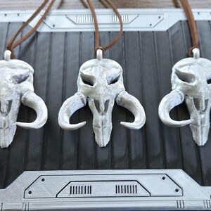 May include: Three identical silver-colored pendants shaped like a stylized creature skull with large curved horns. Each pendant is suspended from a thin brown cord. The skull design features hollow eye sockets and a pointed snout, with a metallic finish.