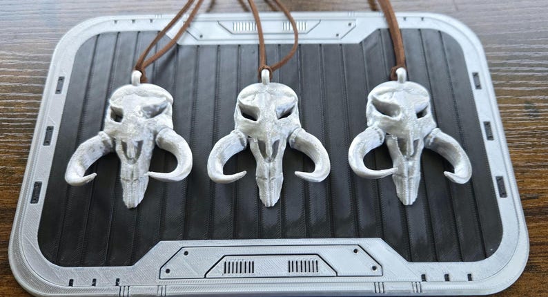 May include: Three identical silver-colored pendants shaped like a stylized creature skull with large curved horns. Each pendant is suspended from a thin brown cord. The skull design features hollow eye sockets and a pointed snout, with a metallic finish.