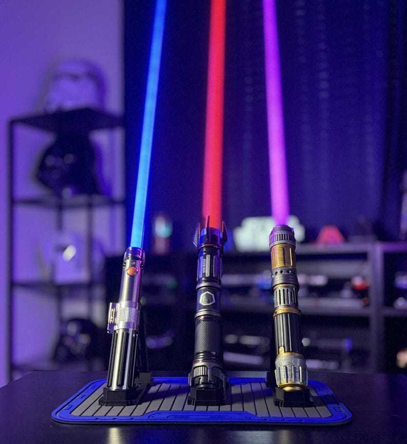 K&ouml;nnte beinhalten: Drei beleuchtete Lichtschwerter mit blauen, roten und violetten Klingen stehen auf einer blau-grauen Matte. Die Griffe sind silber, schwarz und goldfarben. Im Hintergrund sind ein Regal und andere Star Wars-Erinnerungsst&uuml;cke zu sehen.