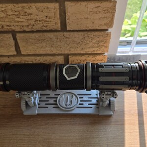 May include: A black lightsaber with red accents and a silver emblem on the side, resting on a gray stand with a Rebel Alliance symbol.
