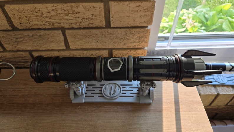 May include: A black lightsaber with red accents and a silver emblem on the side, resting on a gray stand with a Rebel Alliance symbol.