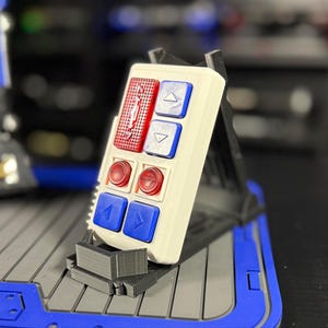 May include: A white rectangular control panel with red and blue buttons. The panel has two rows of buttons, with red buttons on the top and bottom rows and blue buttons in the middle. The panel is mounted on a black stand.