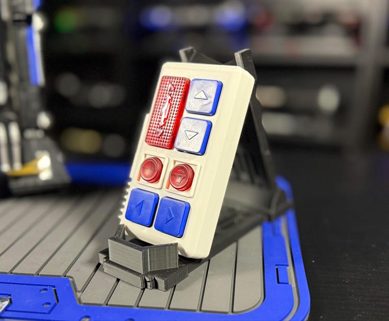 May include: A white rectangular control panel with red and blue buttons. The panel has two rows of buttons, with red buttons on the top and bottom rows and blue buttons in the middle. The panel is mounted on a black stand.
