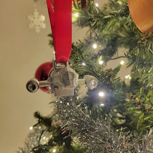 May include: A silver-colored spaceship ornament hanging from a red ribbon on a Christmas tree. The ornament is detailed with a cockpit and wings.