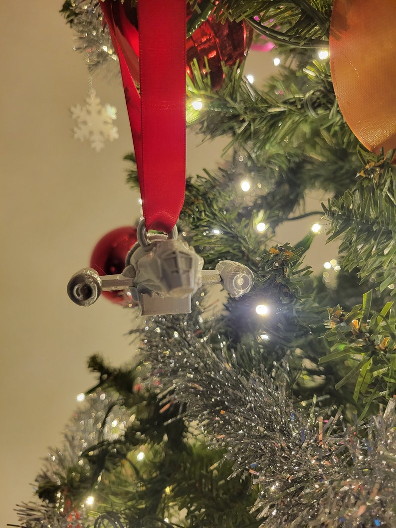 May include: A silver-colored spaceship ornament hanging from a red ribbon on a Christmas tree. The ornament is detailed with a cockpit and wings.