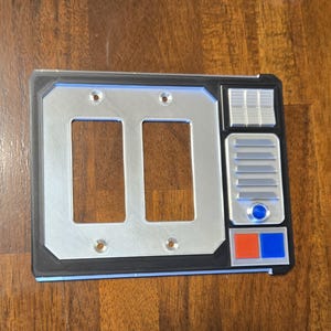 May include: A double light switch cover with a Star Wars themed design. The cover is silver with black trim and features a control panel with buttons and a speaker.