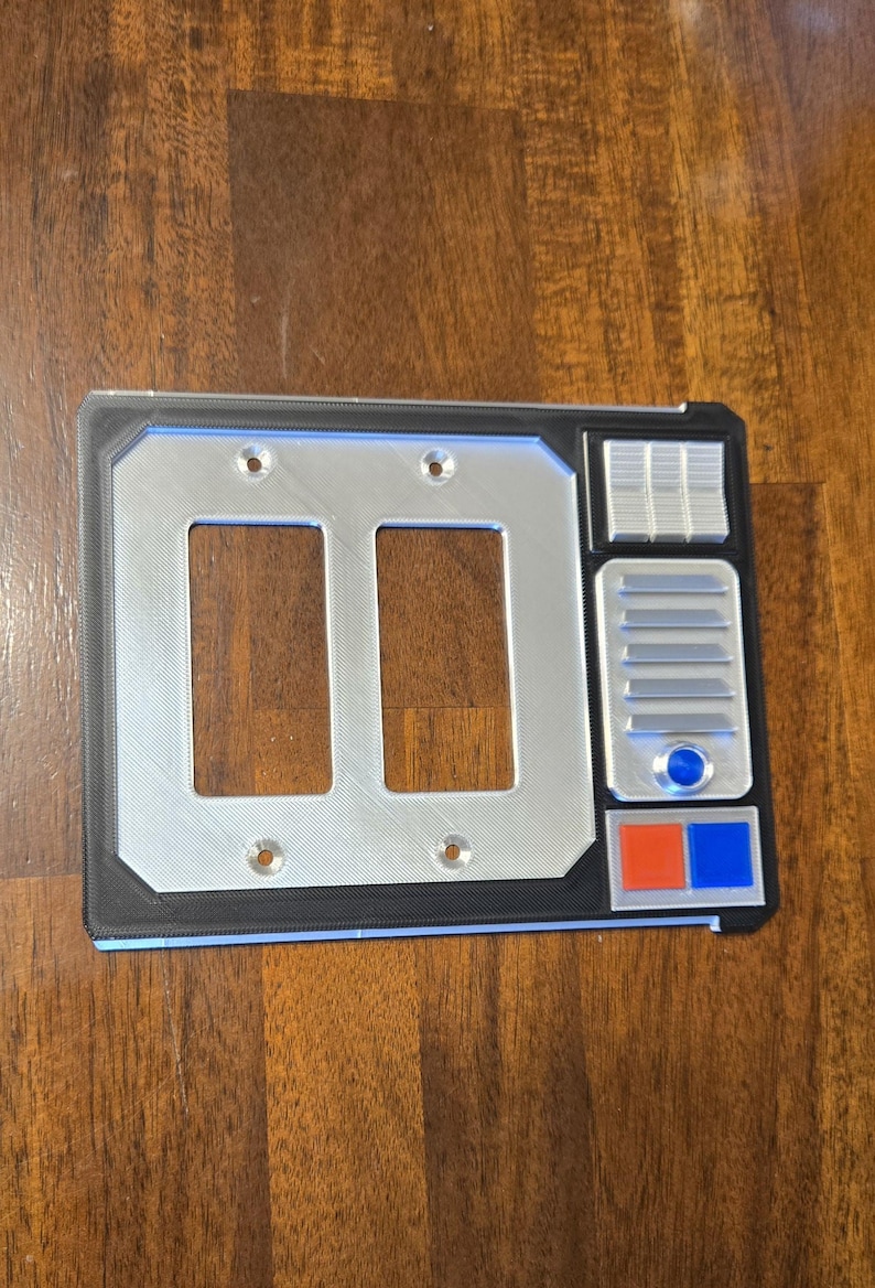 May include: A double light switch cover with a Star Wars themed design. The cover is silver with black trim and features a control panel with buttons and a speaker.