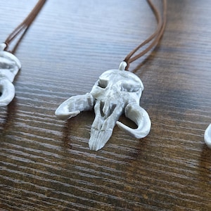 May include: Four pendants shaped like a stylized creature skull with curved horns, finished in a weathered off-white color. Each pendant is suspended from a thin brown cord, featuring a textured surface with visible layered details throughout.