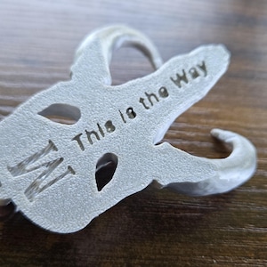 May include: A silver-colored metal pendant shaped like a stylized skull with curved horns, featuring the engraved text "This is the Way" and a letter "M" on its surface, suspended from a thin brown leather cord necklace.