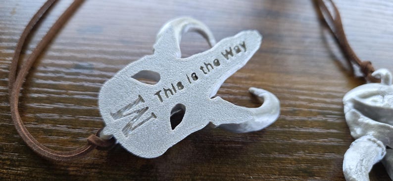 May include: A silver-colored metal pendant shaped like a stylized skull with curved horns, featuring the engraved text "This is the Way" and a letter "M" on its surface, suspended from a thin brown leather cord necklace.