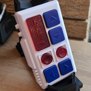 Peut inclure: Un appareil &eacute;lectronique blanc et noir avec des boutons bleus et rouges. L'appareil a une forme rectangulaire avec une s&eacute;rie de boutons et un panneau rouge textur&eacute;. Les boutons sont bleus avec des fl&egrave;ches blanches, et les panneaux rouges ont un motif circulaire.