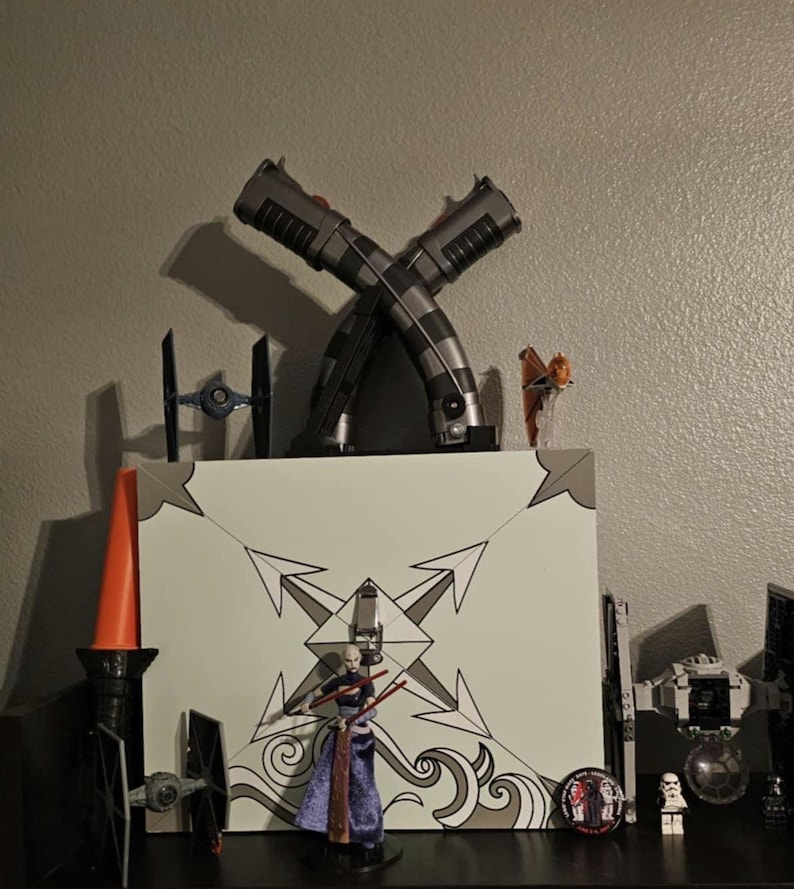 K&ouml;nnte beinhalten: Eine Pr&auml;sentation von Star Wars-Sammlerst&uuml;cken. Zwei gekreuzte Lichtschwerter sind &uuml;ber einer wei&szlig;en Box mit einem geometrischen Design positioniert. Weitere Artikel sind Miniatur-TIE-J&auml;ger, Figuren und ein rot-oranges Lichtschwert.