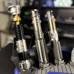 May include: Three metallic lightsabers displayed on a blue and gray stand. The lightsabers have silver, black, and gold accents, with various button and switch details. One lightsaber is partially disassembled, showcasing internal components.