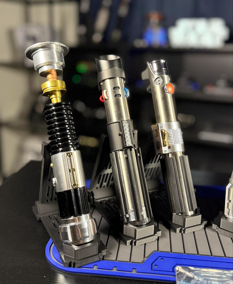 May include: Three metallic lightsabers displayed on a blue and gray stand. The lightsabers have silver, black, and gold accents, with various button and switch details. One lightsaber is partially disassembled, showcasing internal components.