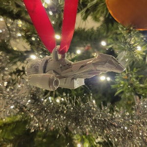 May include: A white 3D printed spaceship ornament hanging from a red ribbon on a Christmas tree.