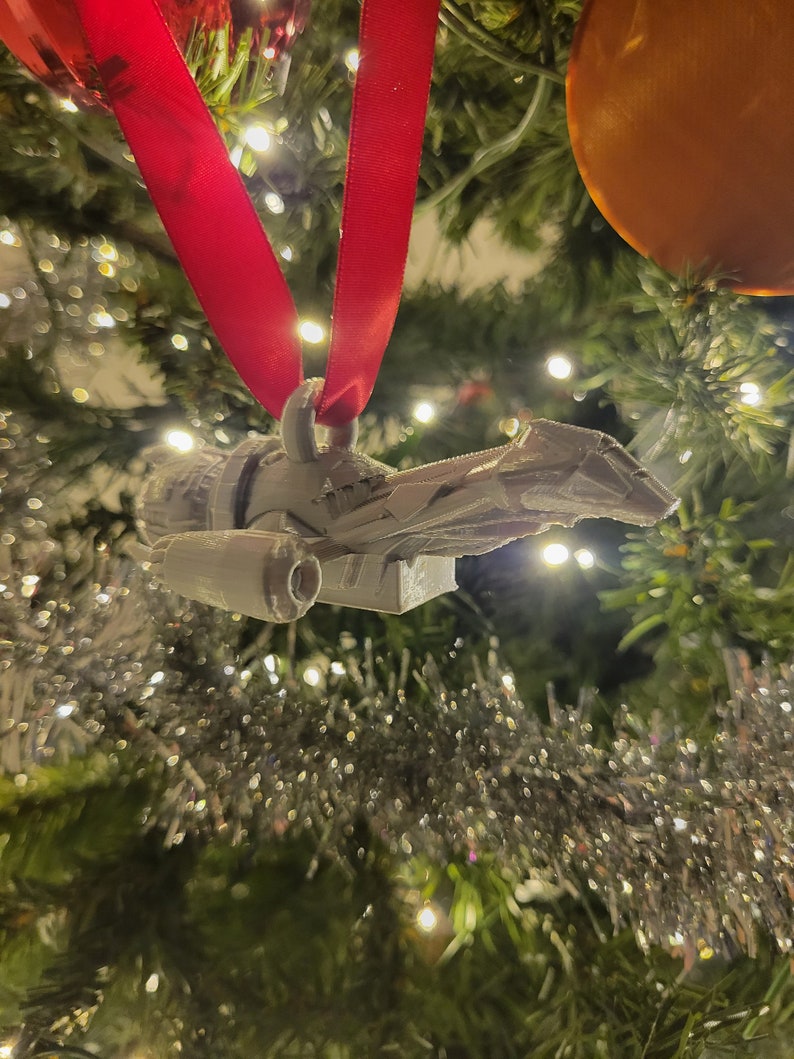 May include: A white 3D printed spaceship ornament hanging from a red ribbon on a Christmas tree.