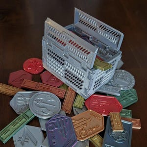 May include: A 3D printed storage box with a lid open, revealing a collection of colorful tokens. The tokens are made of plastic and have various shapes and designs. The box is made of a light gray plastic and has a textured surface.