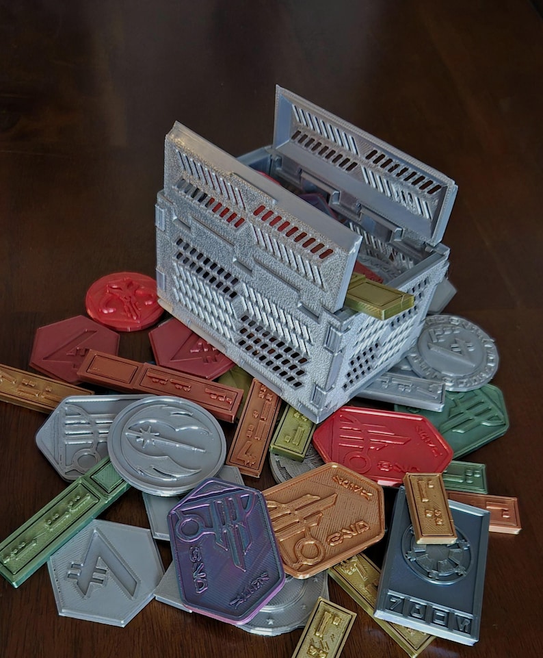 May include: A 3D printed storage box with a lid open, revealing a collection of colorful tokens. The tokens are made of plastic and have various shapes and designs. The box is made of a light gray plastic and has a textured surface.