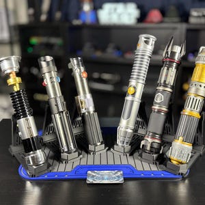 May include: A collection of six detailed lightsabers in various designs, displayed on a blue and gray stand. The lightsabers feature metallic silver, black, and gold accents, with intricate detailing and varying handle designs.