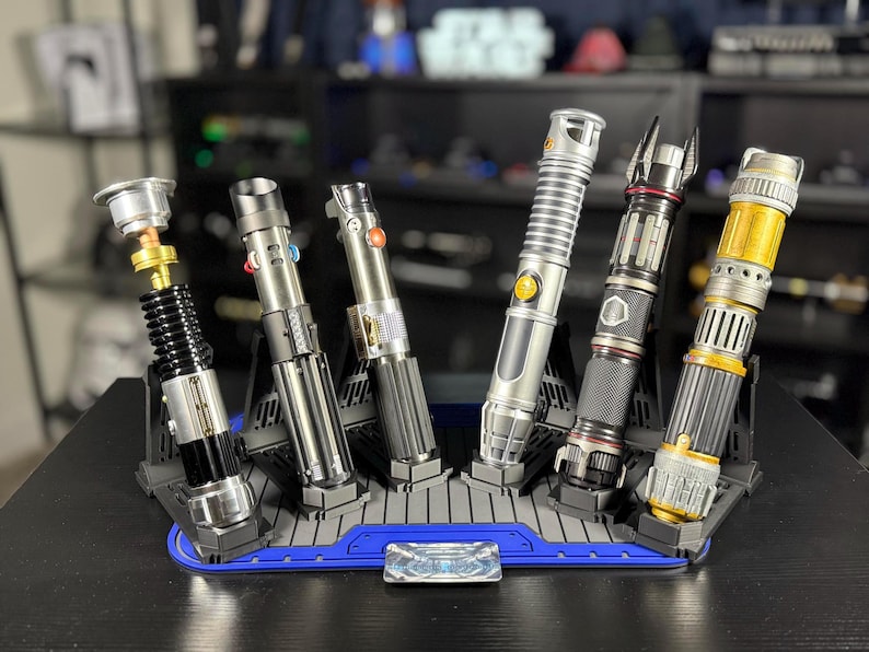 May include: A collection of six detailed lightsabers in various designs, displayed on a blue and gray stand. The lightsabers feature metallic silver, black, and gold accents, with intricate detailing and varying handle designs.