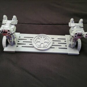 May include: A 3D printed display stand for two Star Wars AT-ST walkers. The stand is gray and features a raised Imperial symbol in the center.