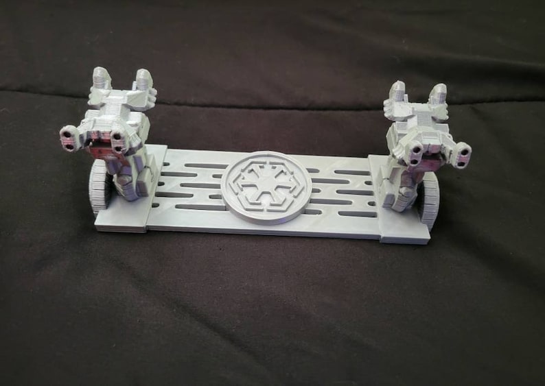 May include: A 3D printed display stand for two Star Wars AT-ST walkers. The stand is gray and features a raised Imperial symbol in the center.