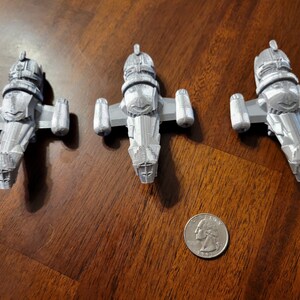 May include: Three silver-colored miniature spaceships, resembling the Serenity from the TV show Firefly, are arranged on a wooden surface. A US quarter is included for scale.
