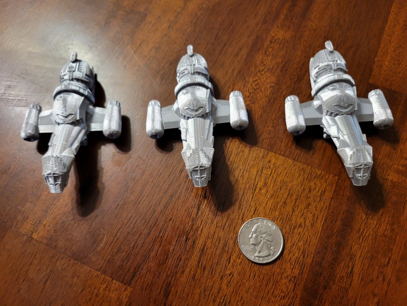 May include: Three silver-colored miniature spaceships, resembling the Serenity from the TV show Firefly, are arranged on a wooden surface. A US quarter is included for scale.