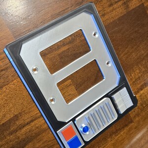 May include: A silver and black wall plate with two rectangular openings and a row of buttons. The buttons are blue, orange, and silver.