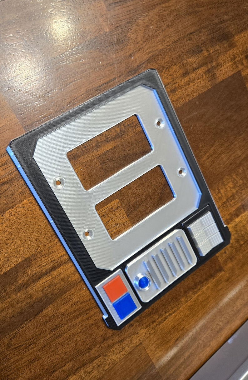 May include: A silver and black wall plate with two rectangular openings and a row of buttons. The buttons are blue, orange, and silver.