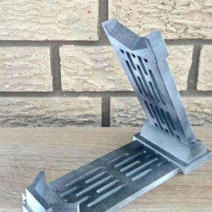 Op de afbeelding: Een grijze 3D-geprinte telefoonstandaard met een geometrisch ontwerp. De standaard is open en klaar om een telefoon te houden.